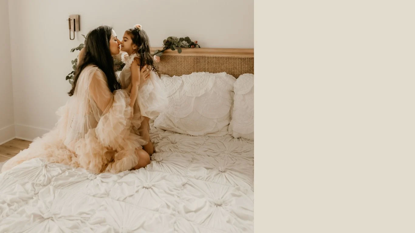 Mother and daughter in soft tulle dresses sharing a kiss on a styled bed in a natural light bedroom
