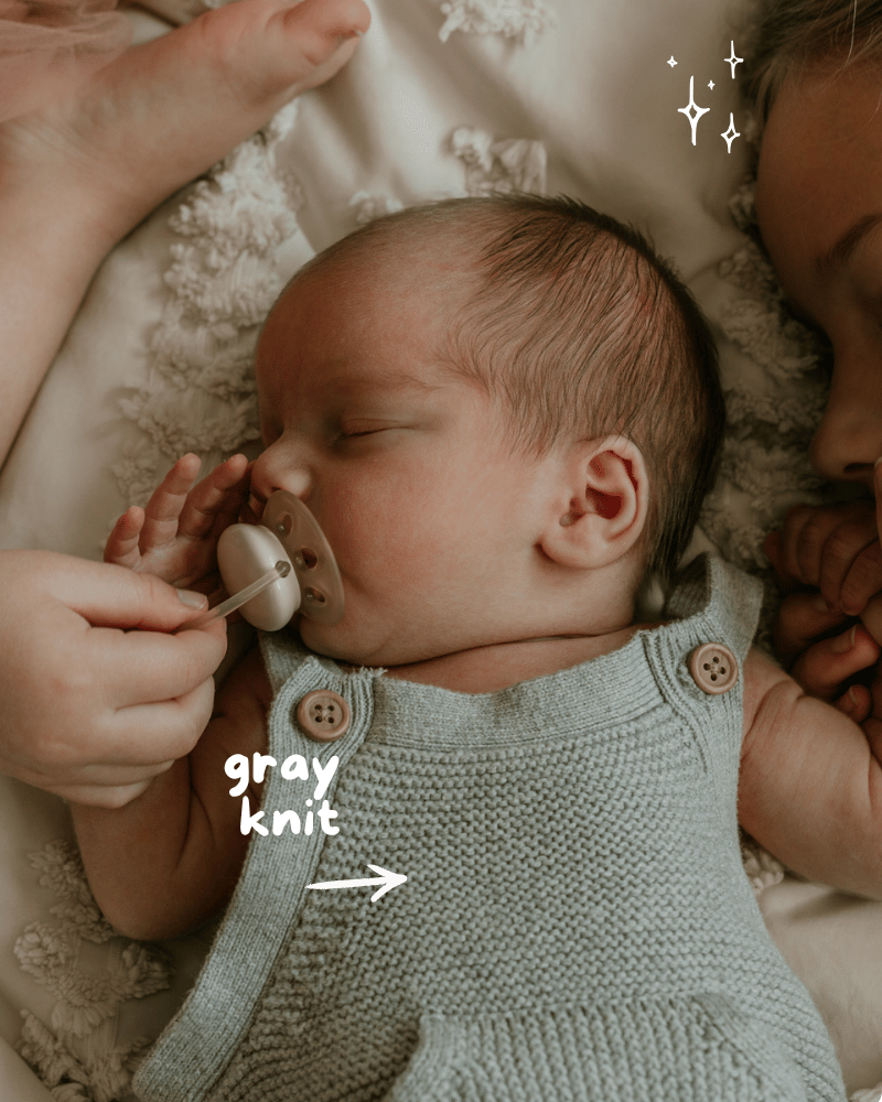 Newborn in gray knit romper with pacifier, cuddled beside sibling on textured white blanket