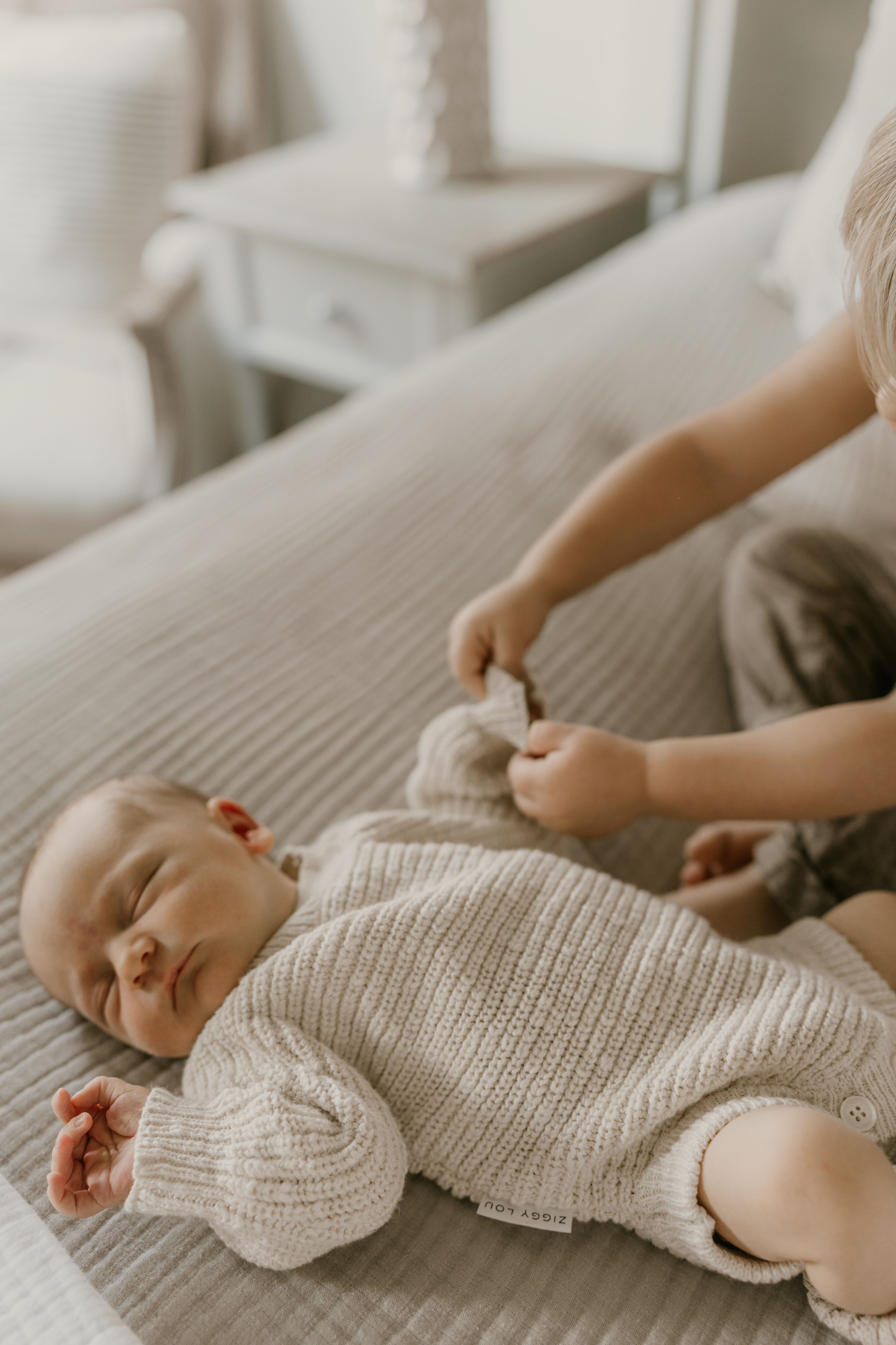 Older sibling gently holding newborn’s sleeve during cozy in-home newborn session on a bed