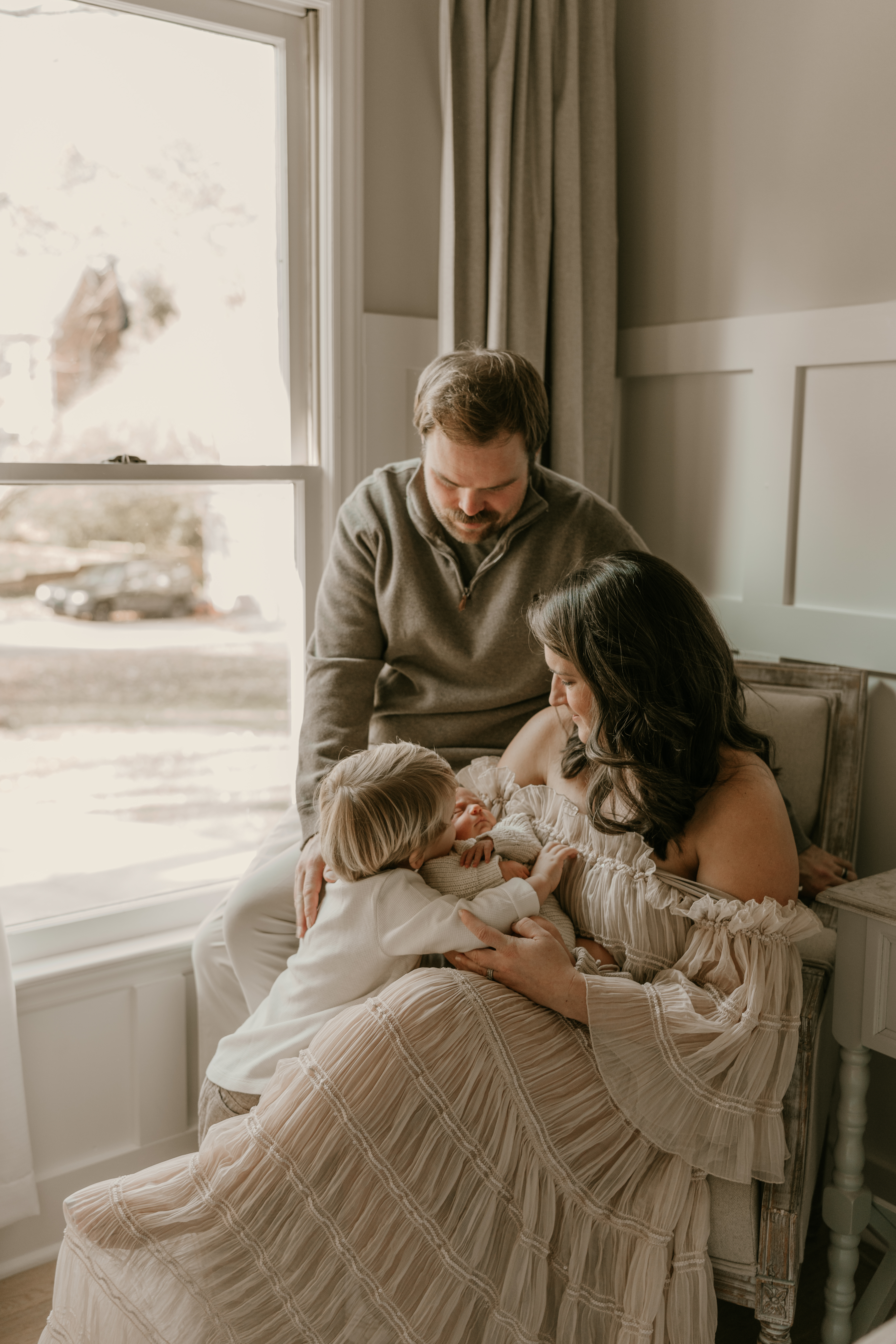 Mother holding newborn while toddler embraces baby, father leaning over with a loving gaze, natural light pouring through a window