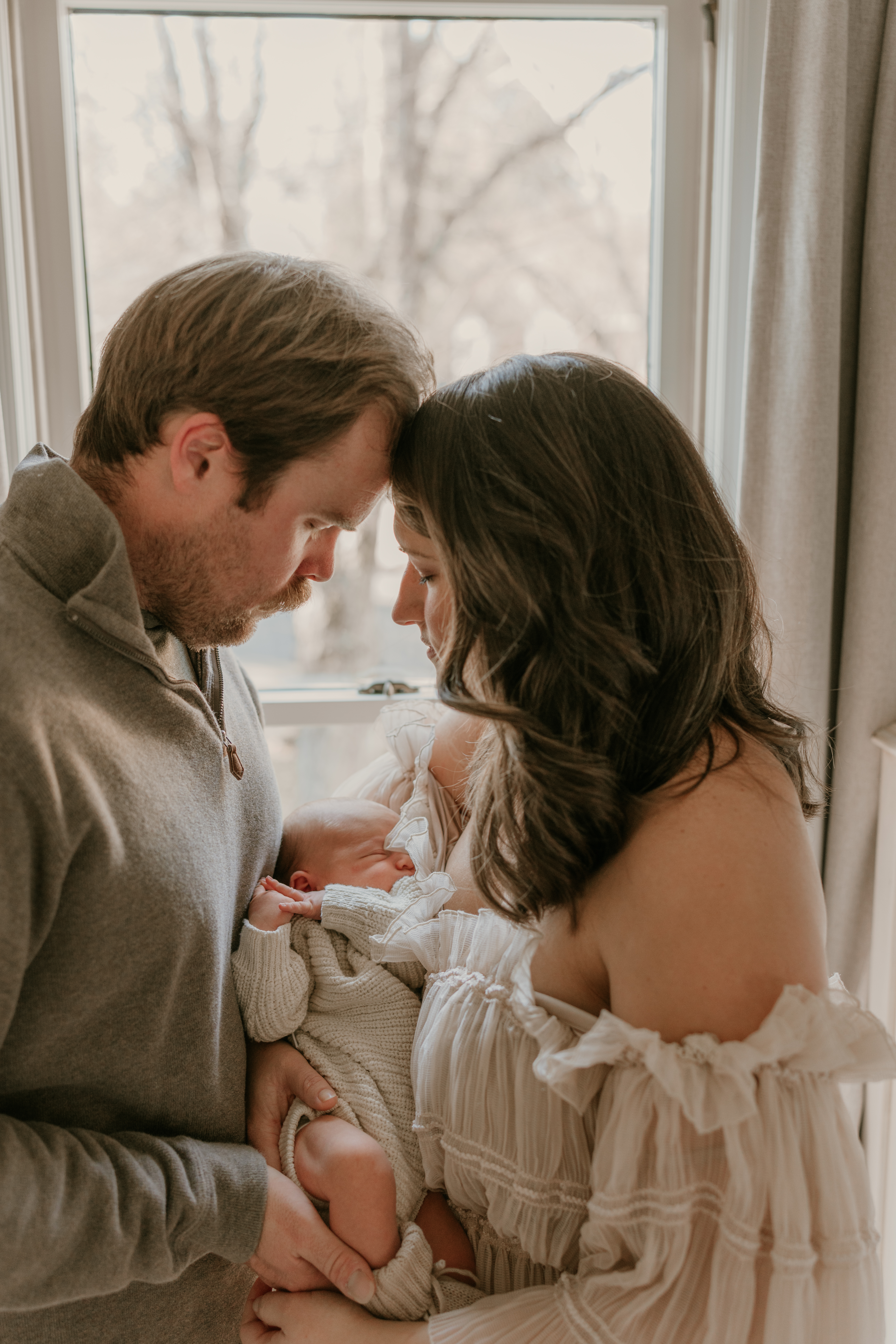Mother and father holding newborn baby while gently touching foreheads near a window