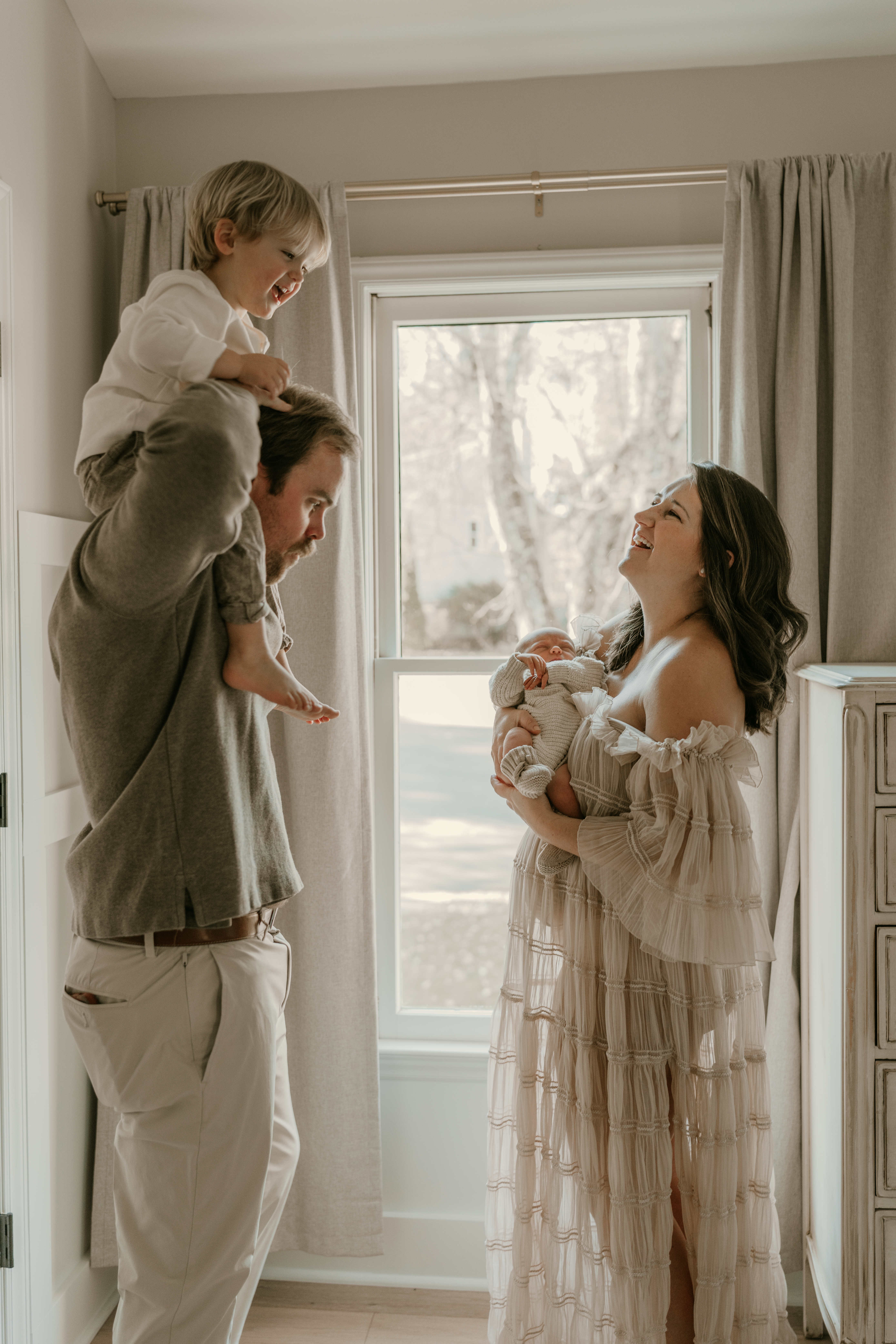 Smiling mother holding newborn as father lifts toddler on shoulders near a bright window