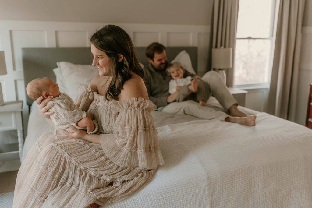 Mother holding newborn while father and toddler play on bed during family lifestyle session