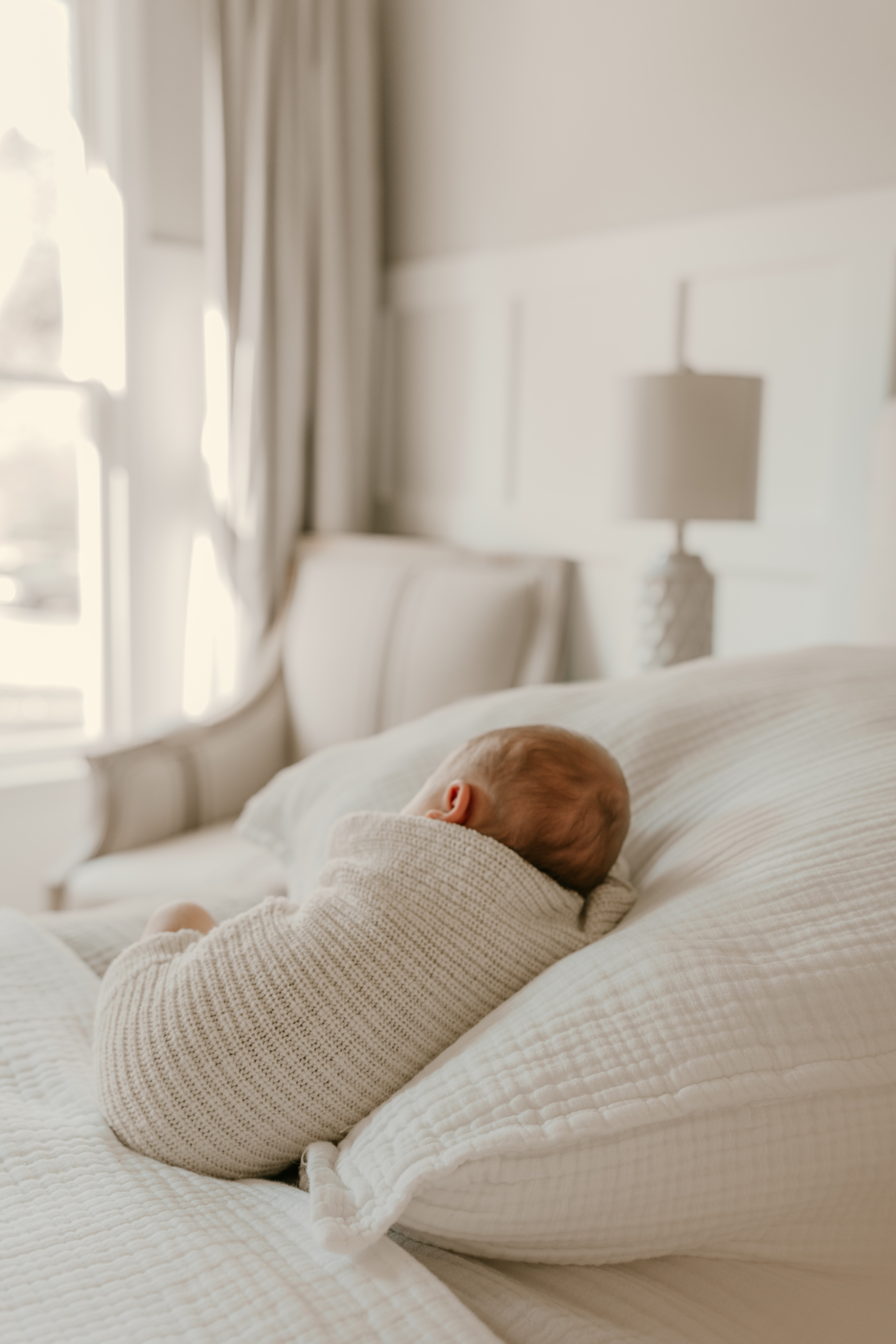 Newborn baby in knit outfit lying on white bed near window with soft morning light