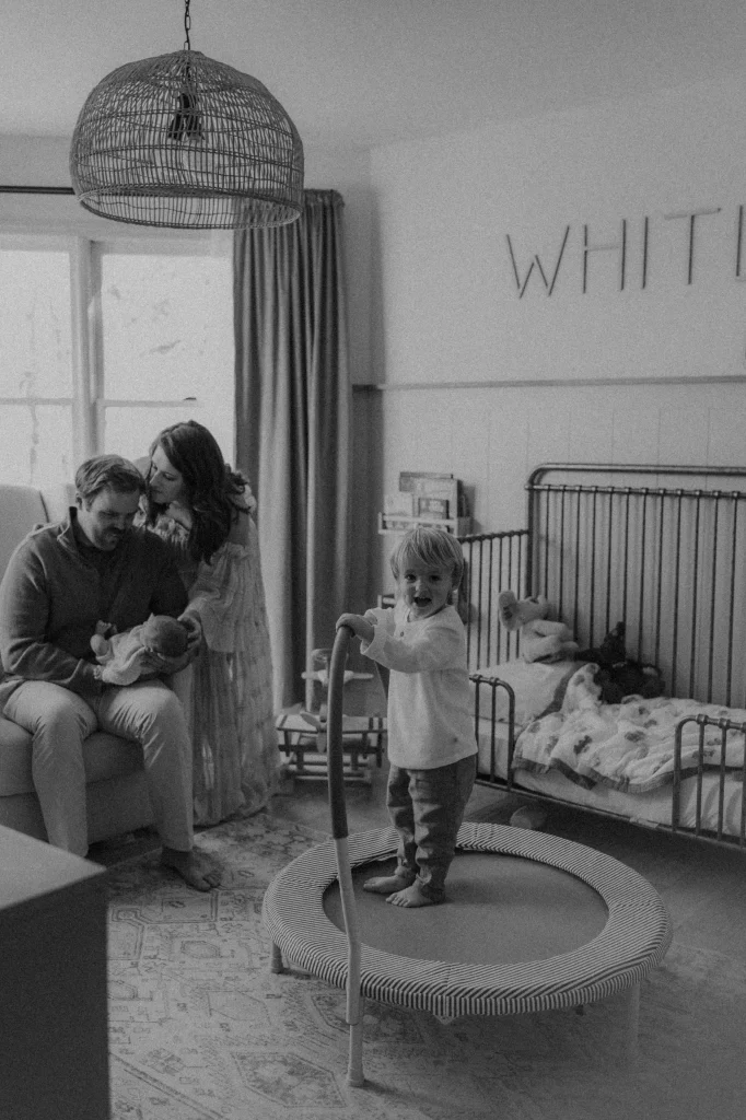 Family in nursery with toddler on trampoline and parents admiring newborn