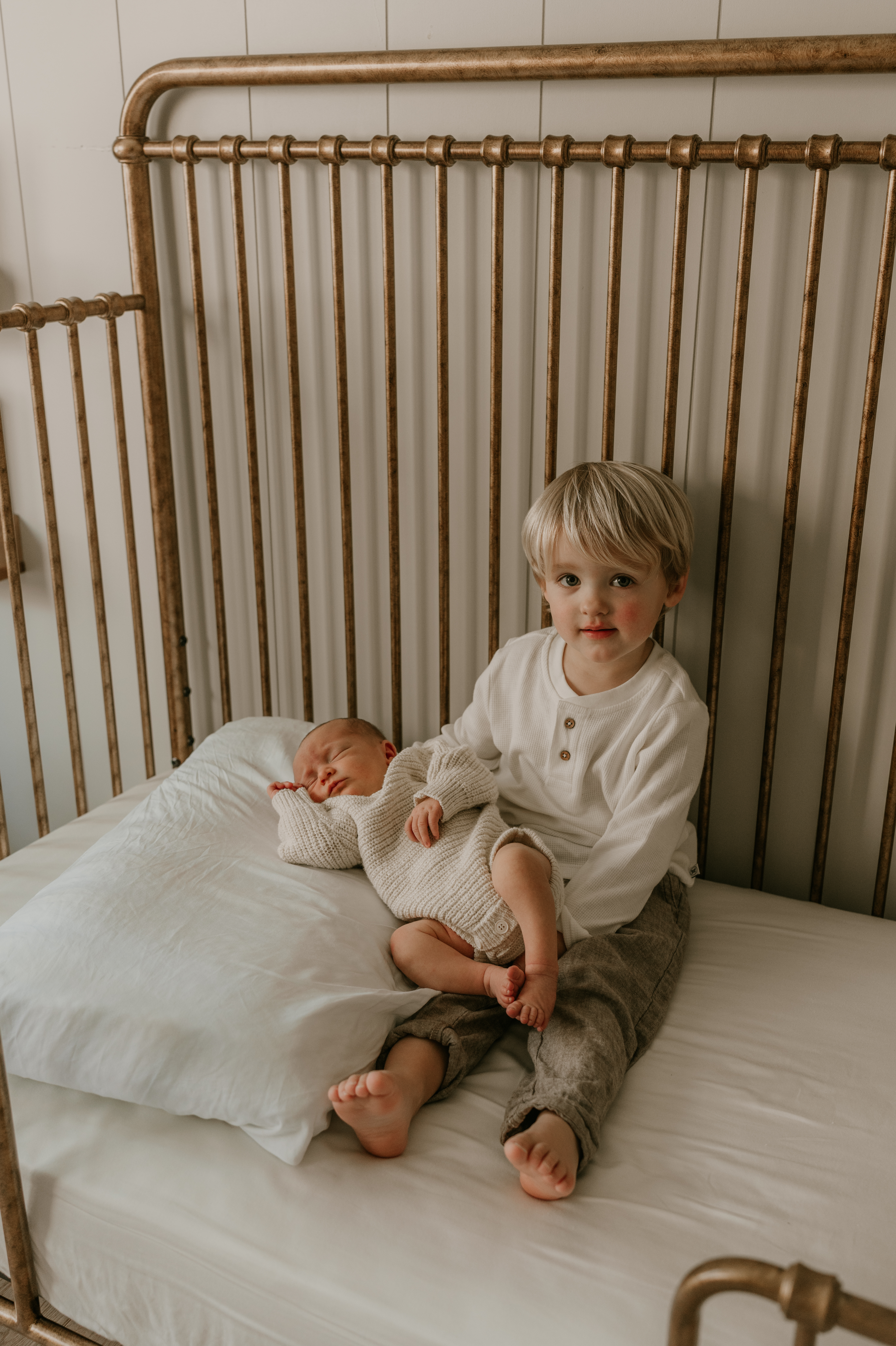 Toddler sitting in crib holding newborn sibling in matching neutral tones