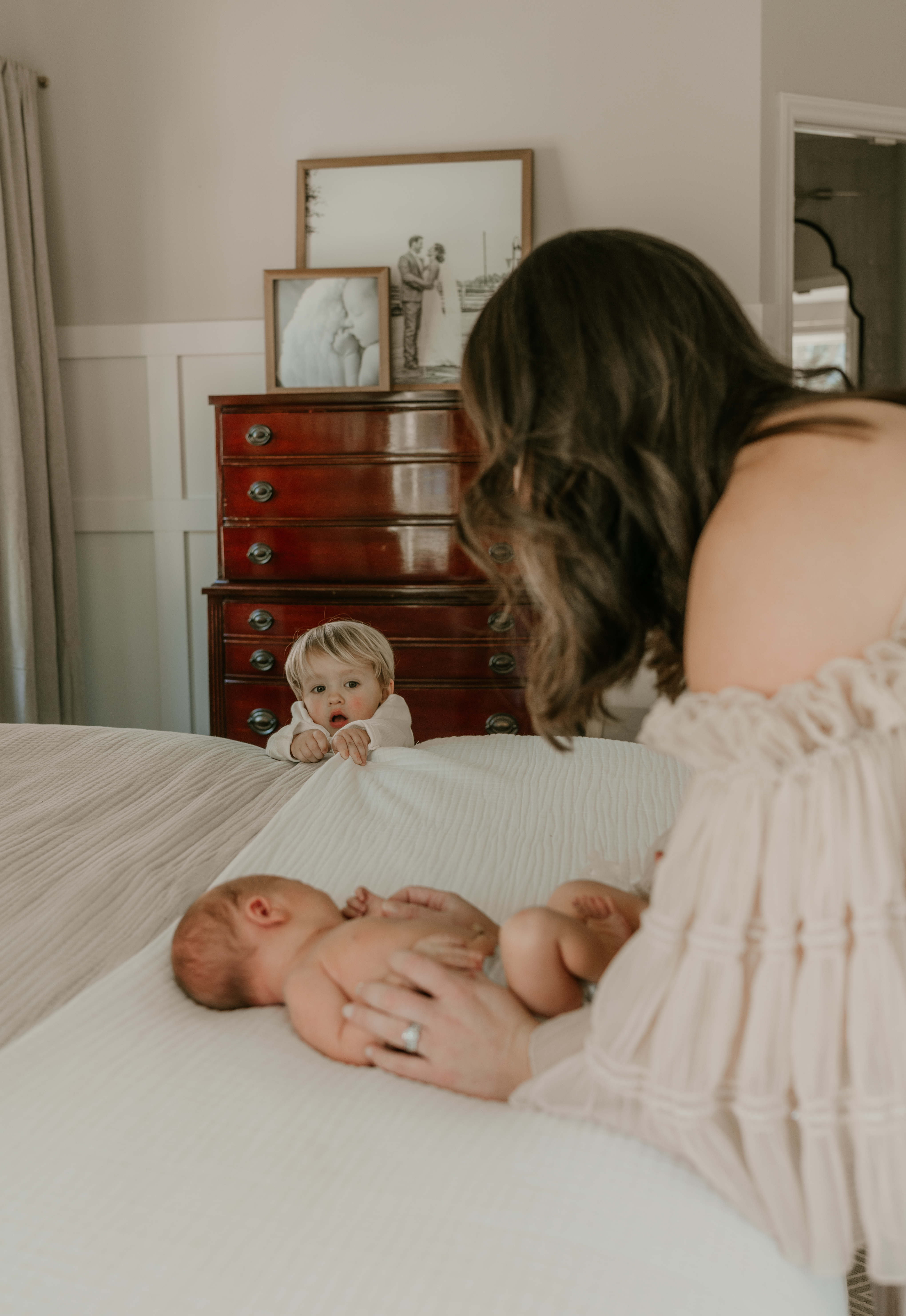 Toddler peeking over the bed at newborn lying near mother’s hand