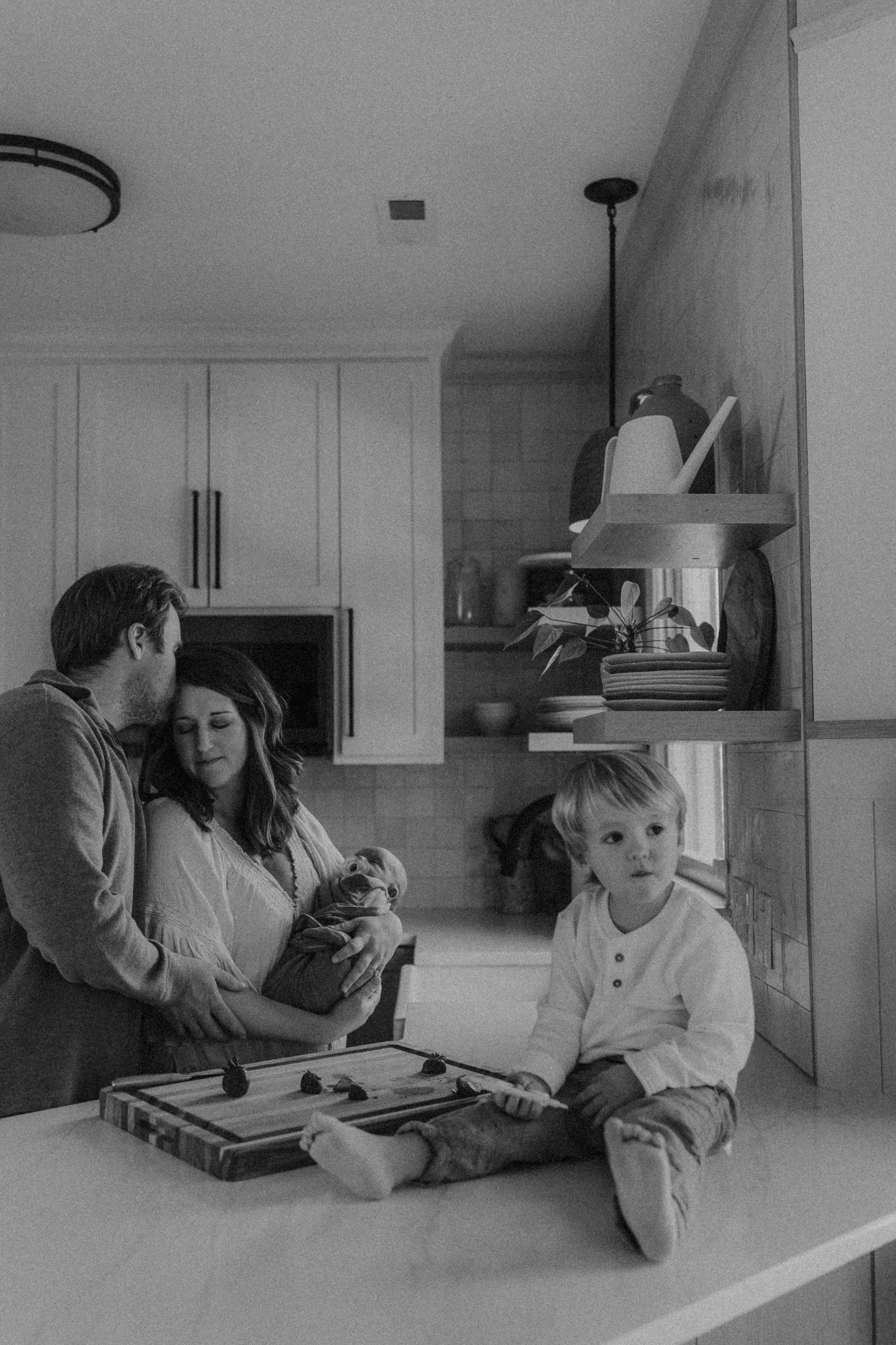 Family moment in kitchen with toddler eating and parents snuggling newborn