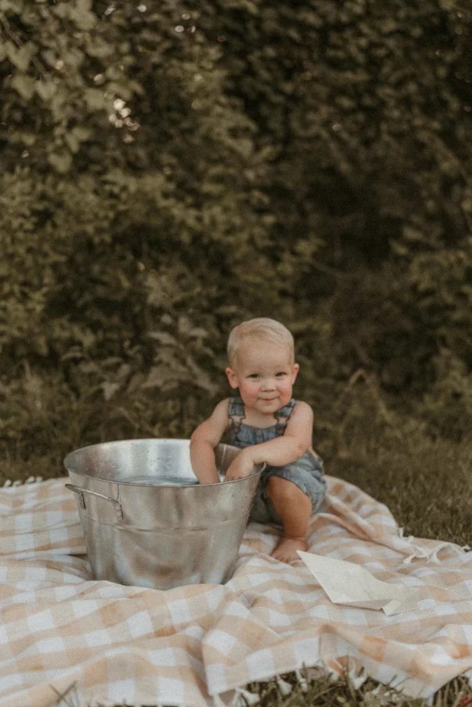 Toddler in denim romper playing with a metal water bucket on a gingham blanket during a summer photoshoot