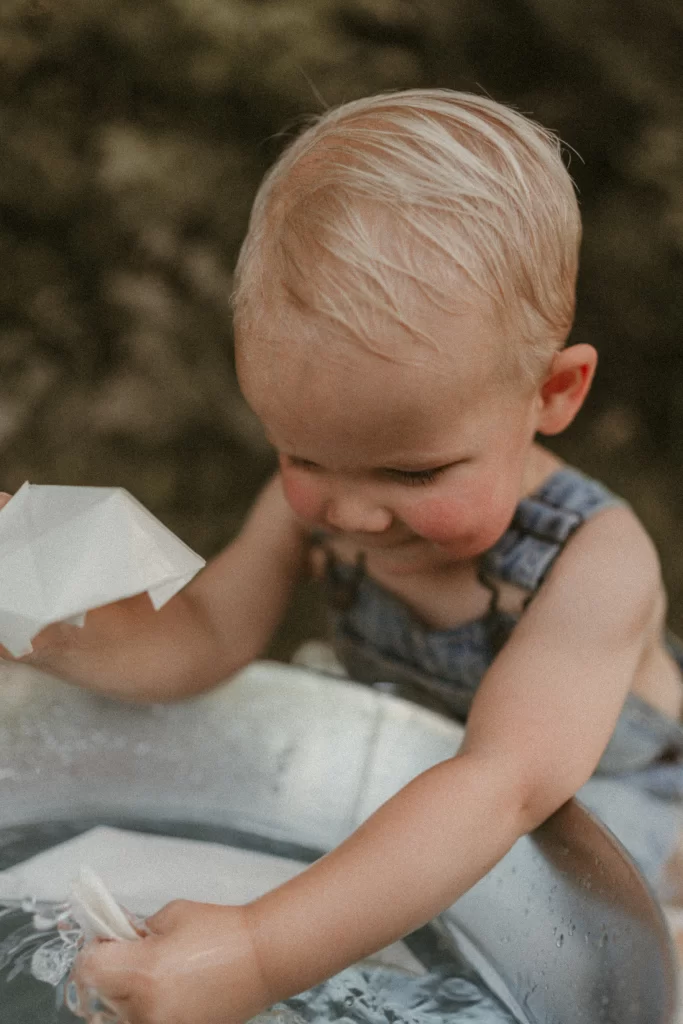 Wet-haired toddler smiling while playing with paper boats in a water-filled metal bucket during a summer outdoor activity