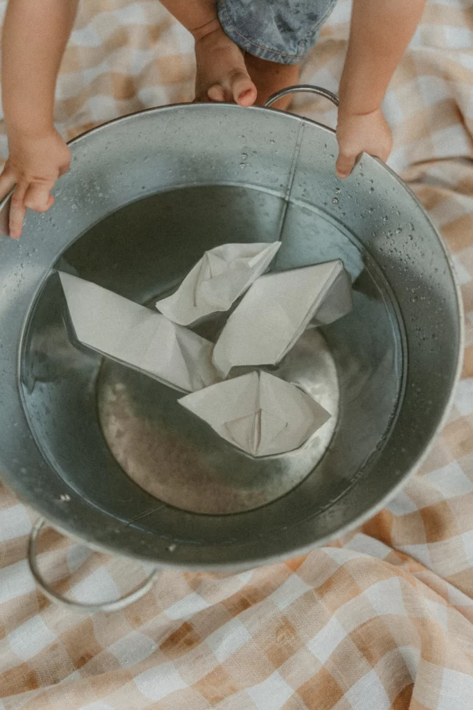 A candid summer moment of a toddler cooling off with a metal bucket of water, captured during an outdoor photo session with warm tones and natural textures perfect for seasonal storytelling.