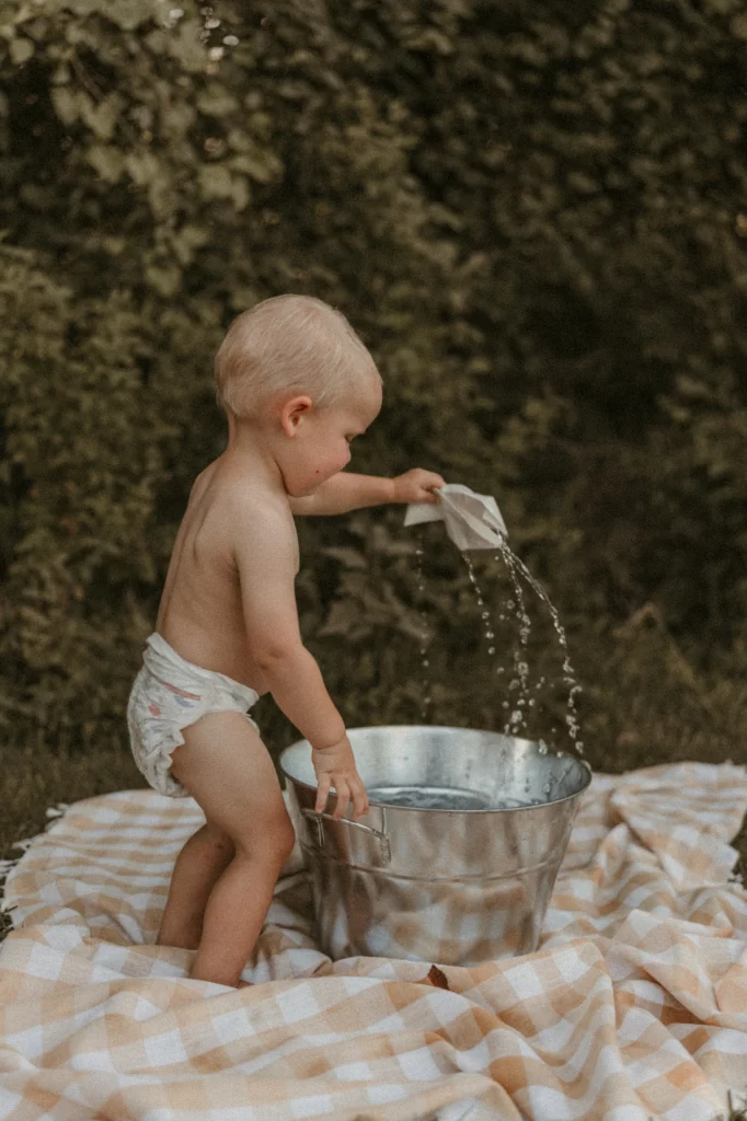 Toddler in a diaper playing with water in a metal bucket on a gingham blanket during a summer outdoor photoshoot