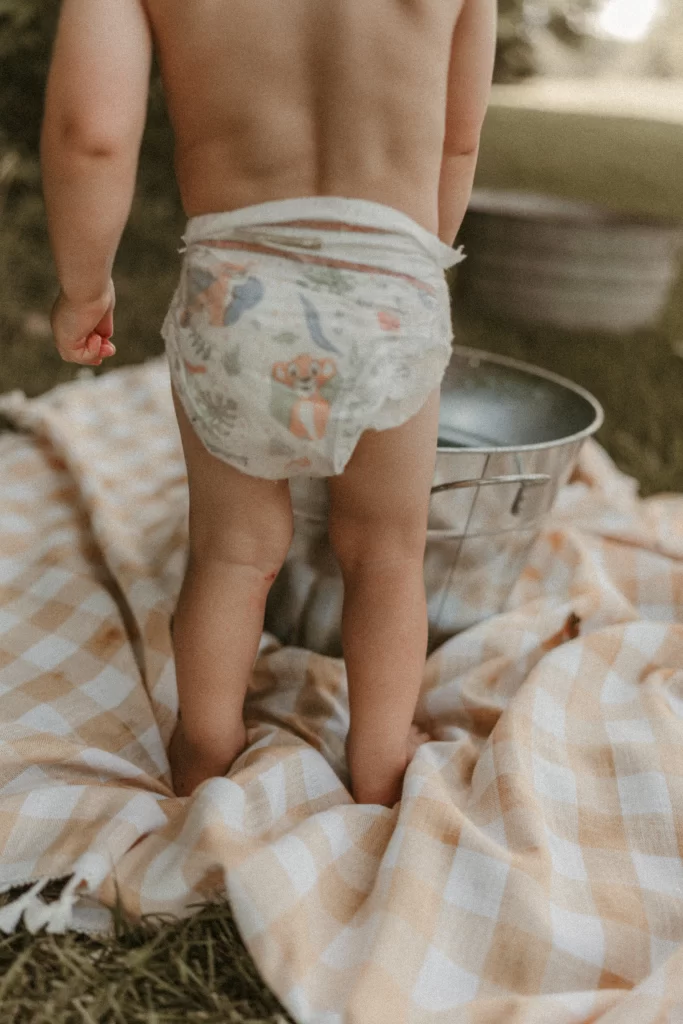 Toddler standing barefoot in a diaper on a gingham blanket next to a metal water bucket during a summer outdoor session