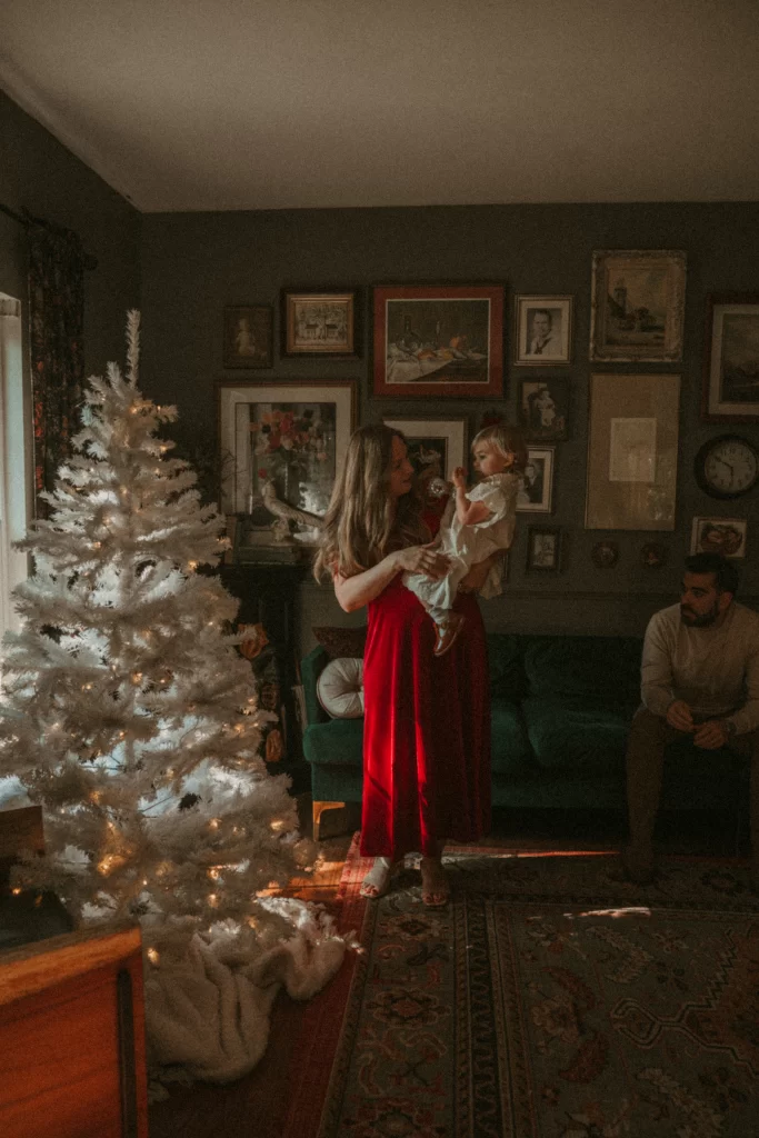 Mother holding toddler by a white Christmas tree with glowing lights, father sitting on sofa in cozy living room