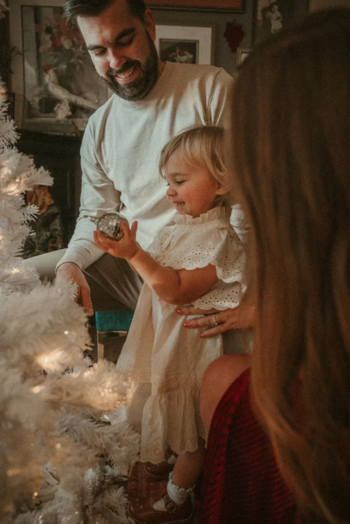 Father smiling while toddler decorates white Christmas tree with ornament, mother nearby