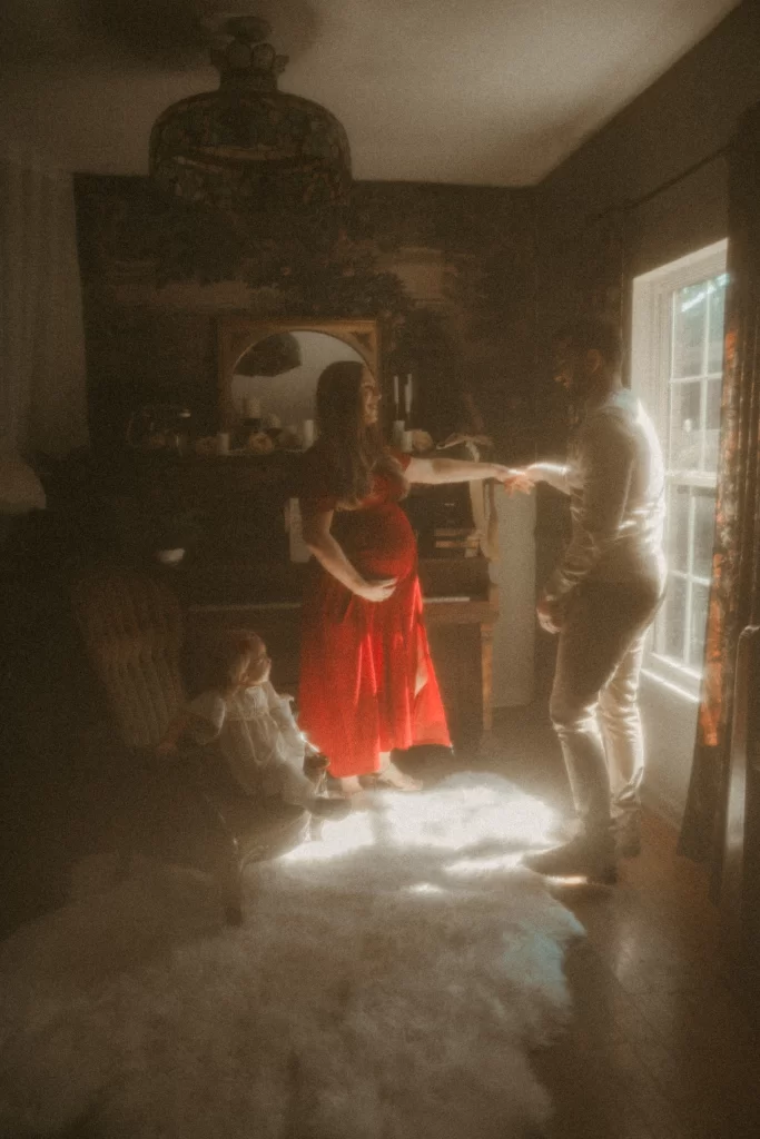 Pregnant mother in red dress holding hands with husband while toddler watches by the piano