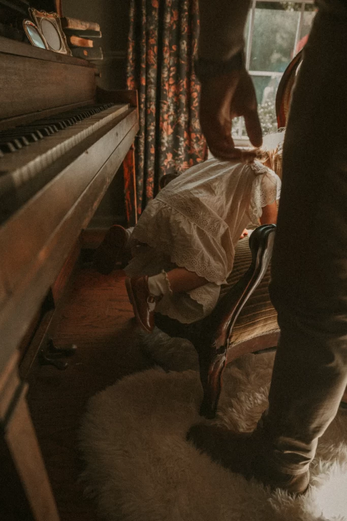 Toddler in white lace dress climbing on chair near piano during family Christmas session