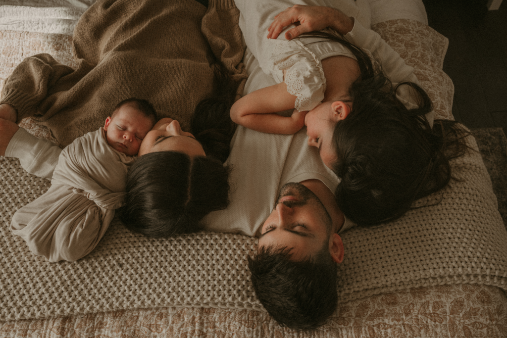 Parents lying on a bed with their newborn baby and toddler, sharing a quiet, intimate family moment
