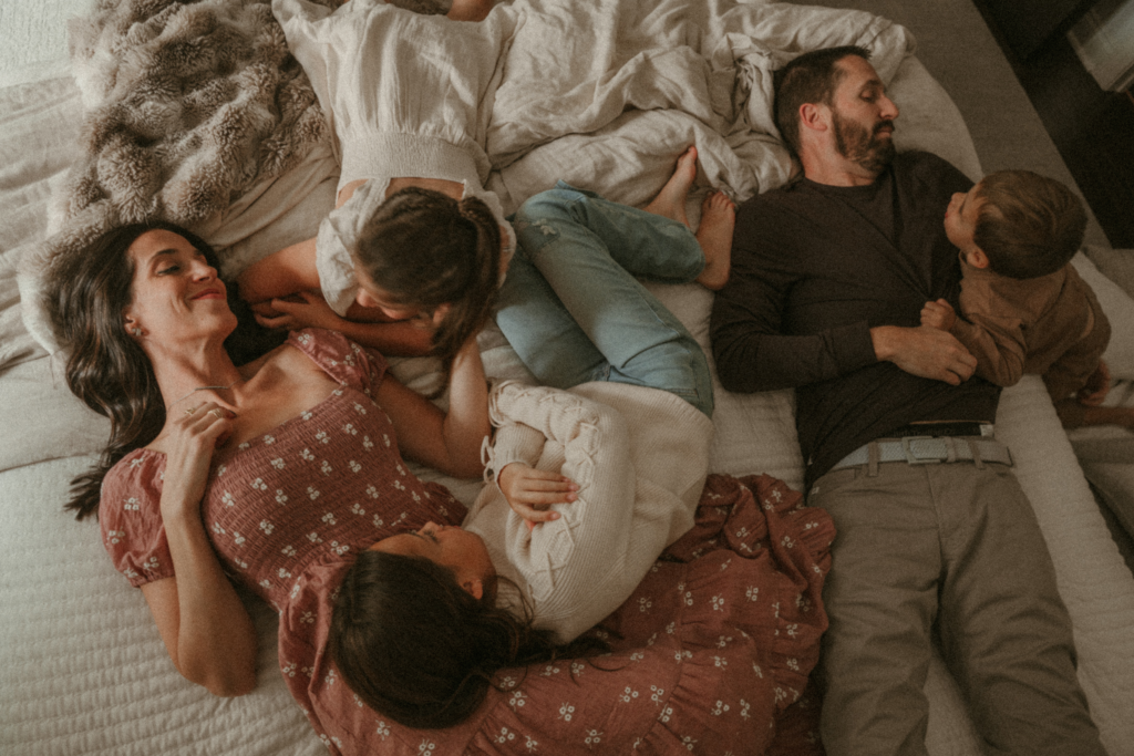 Family lying together on a bed during an in-home lifestyle photo session, capturing candid connection and calm moments