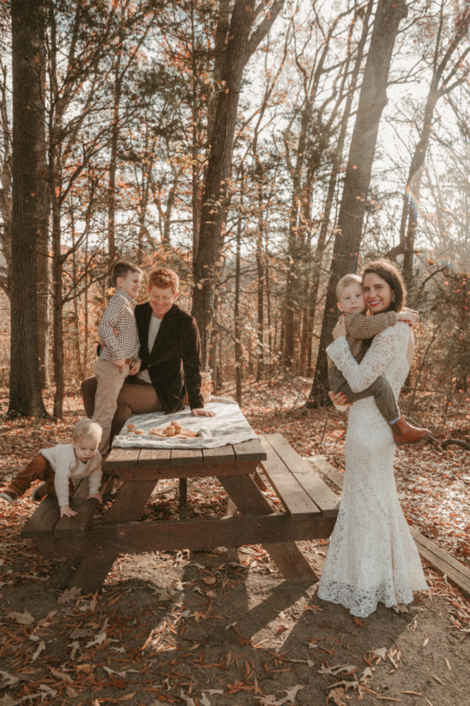 Fall family photo in the woods with parents and young children gathered around a picnic table with warm sunlight
