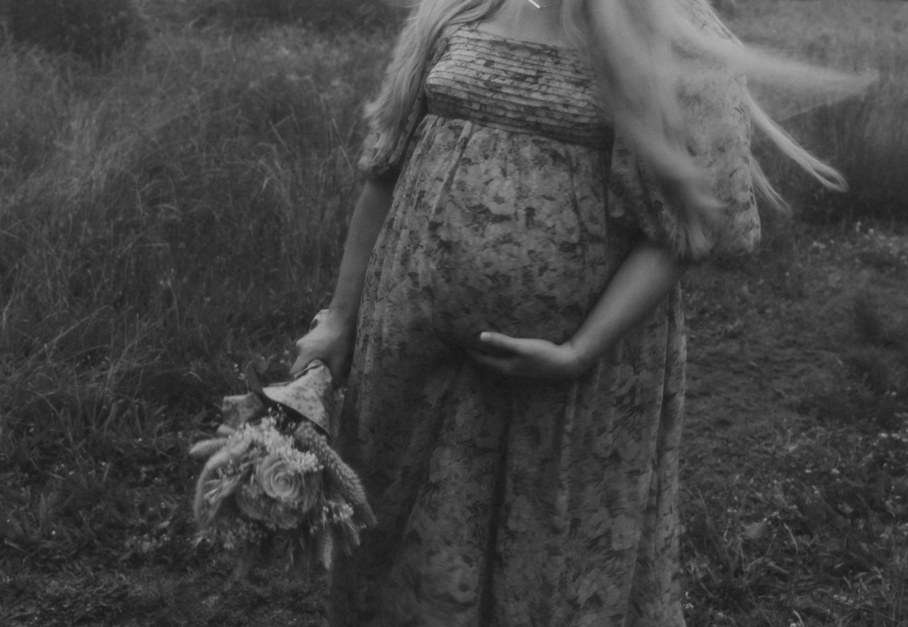 Black and white maternity photo of an expectant mother holding flowers in a grassy field with soft movement