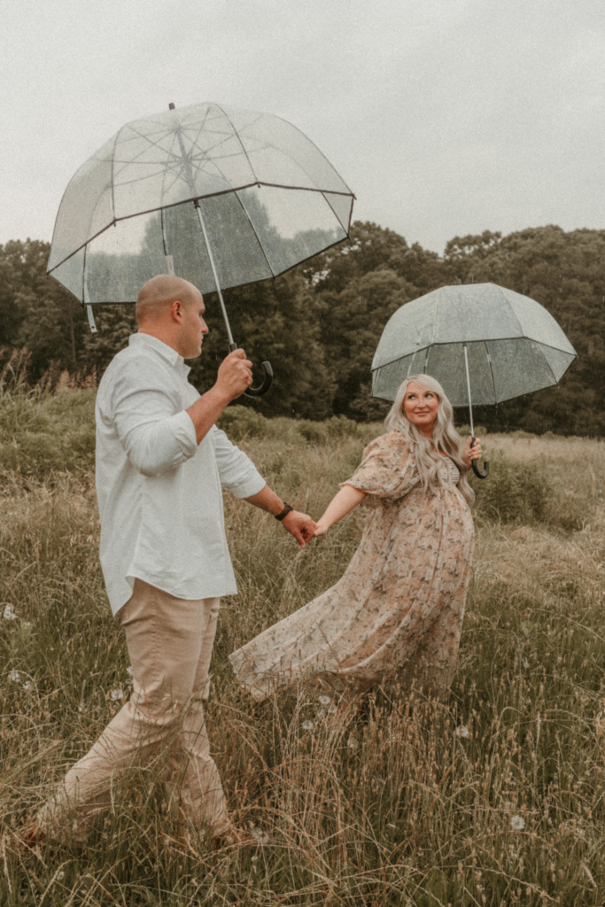 Pregnant couple walking through a field holding hands under clear umbrellas during a moody maternity photo session