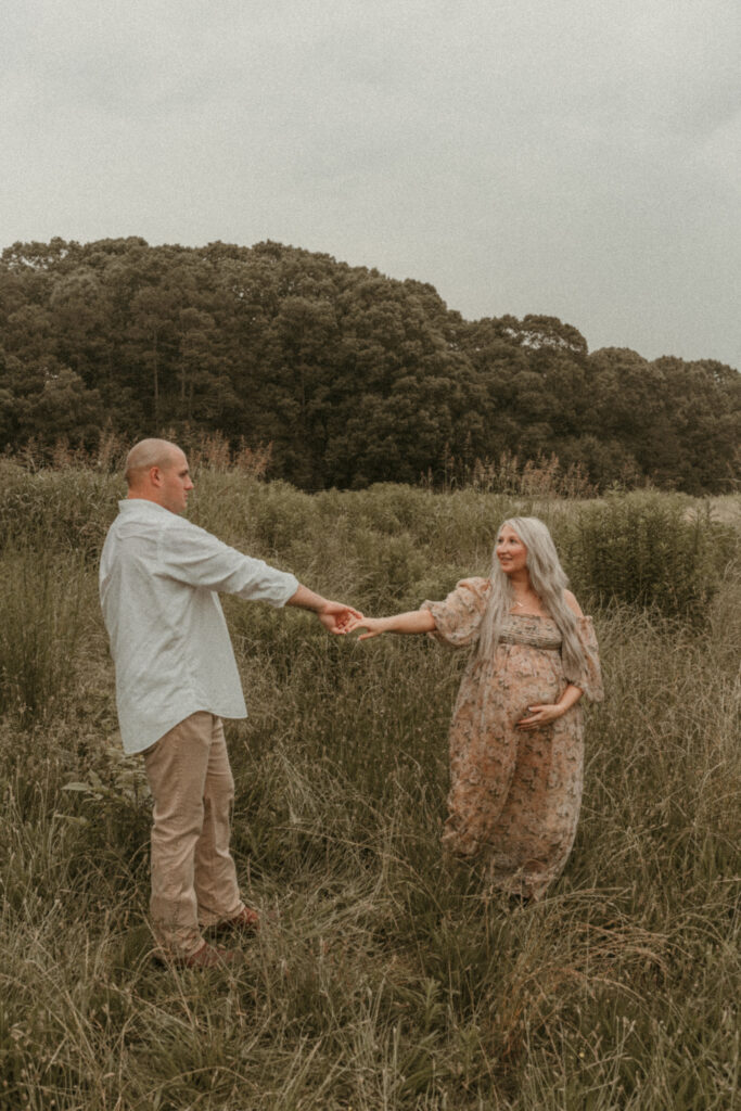 A cinematic outdoor maternity photo featuring an expecting couple dancing through tall grass.. This image highlights connection, movement, and storytelling in a natural, documentary-style maternity session.
